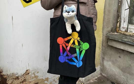 Torba z logo Fediwersum, z której wystaje maskotka pieska. W tle Łódź.